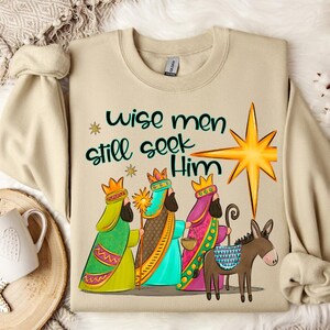 Puede incluir: Una sudadera beige con un dise&ntilde;o navide&ntilde;o. El dise&ntilde;o presenta a los tres reyes magos, un burro y una estrella. El texto en la sudadera dice "Wise men still seek Him".