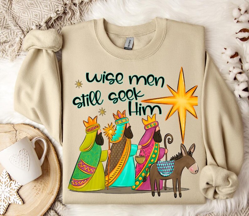 Puede incluir: Una sudadera beige con un dise&ntilde;o navide&ntilde;o. El dise&ntilde;o presenta a los tres reyes magos, un burro y una estrella. El texto en la sudadera dice "Wise men still seek Him".