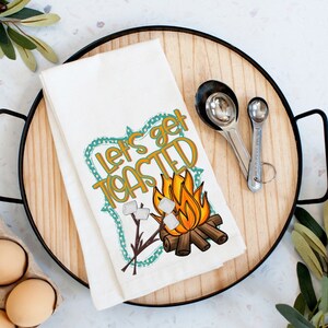 May include: White kitchen towel with a campfire scene and the text "Let's get toasted". The campfire is orange and yellow with white marshmallows on sticks. The text is in a teal and orange frame.
