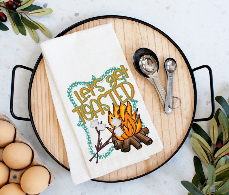 May include: White kitchen towel with a campfire scene and the text "Let's get toasted". The campfire is orange and yellow with white marshmallows on sticks. The text is in a teal and orange frame.