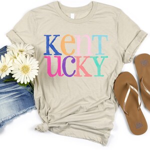 May include: A beige t-shirt with the word "Kentucky" printed in colorful letters. The shirt is styled with denim shorts, white flowers, and brown flip-flops, creating a casual, summery look.