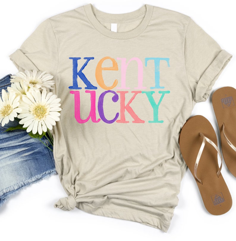 May include: A beige t-shirt with the word "Kentucky" printed in colorful letters. The shirt is styled with denim shorts, white flowers, and brown flip-flops, creating a casual, summery look.