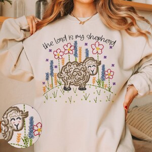 May include: Beige sweatshirt with embroidered design. The design features a sheep, flowers, and the text "the lord is my shepherd". The embroidery includes shades of brown, pink, purple, green, and yellow.