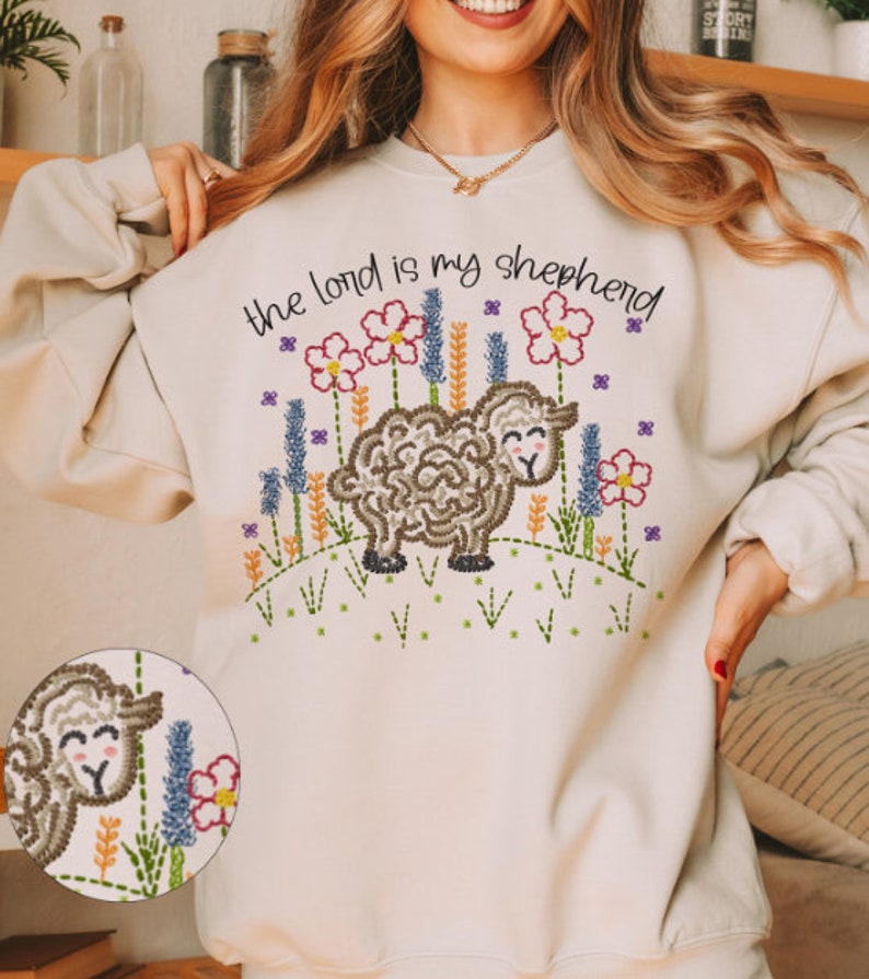 May include: Beige sweatshirt with embroidered design. The design features a sheep, flowers, and the text "the lord is my shepherd". The embroidery includes shades of brown, pink, purple, green, and yellow.