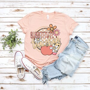 K&ouml;nnte beinhalten: Ein pfirsichfarbenes T-Shirt mit dem Text "Substitute Teacher" in pfirsichfarbener und gelber Schrift. Der Text ist von einem karierten Kreis mit einem Pfirsich-Apfel und Blumen umgeben.