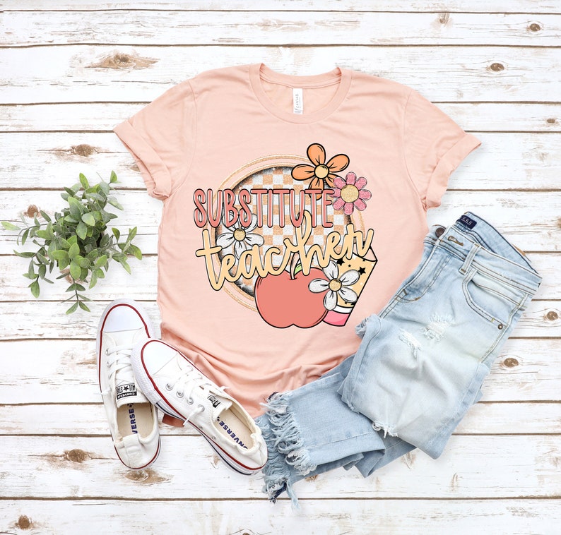 K&ouml;nnte beinhalten: Ein pfirsichfarbenes T-Shirt mit dem Text "Substitute Teacher" in pfirsichfarbener und gelber Schrift. Der Text ist von einem karierten Kreis mit einem Pfirsich-Apfel und Blumen umgeben.