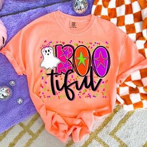 May include: A coral-colored t-shirt with a graphic design that says "BOOtiful". The design includes a white ghost with a pink bow, pink and green confetti, and the word "BOO" in pink, orange, and purple.