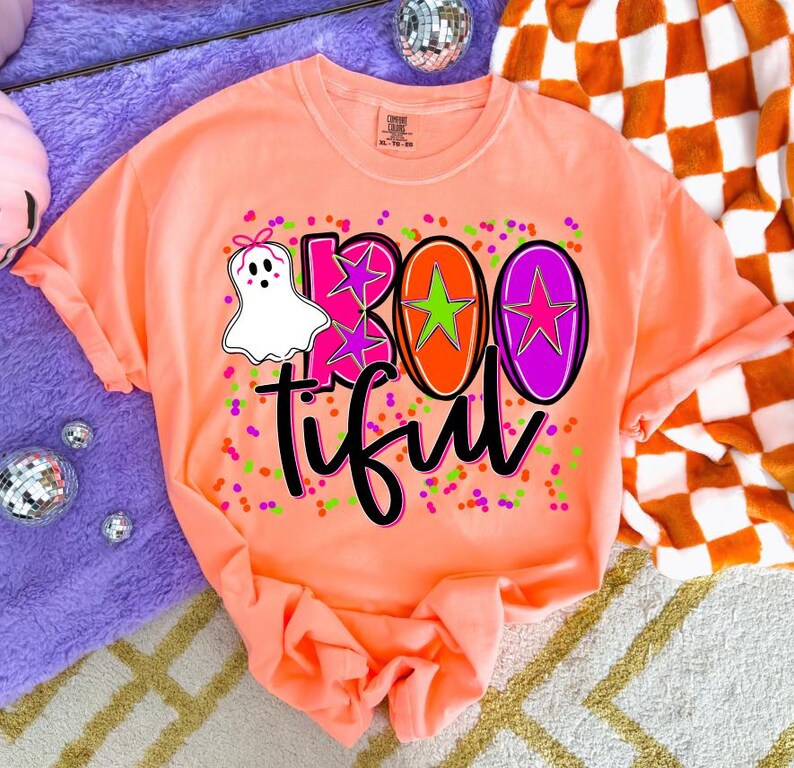 May include: A coral-colored t-shirt with a graphic design that says "BOOtiful". The design includes a white ghost with a pink bow, pink and green confetti, and the word "BOO" in pink, orange, and purple.