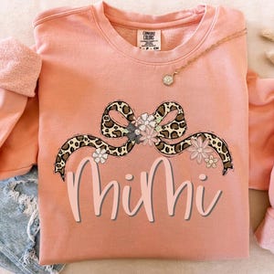 May include: A peach-colored sweatshirt with a leopard print bow and floral accents. The word "Mimi" is written in pink below the bow.