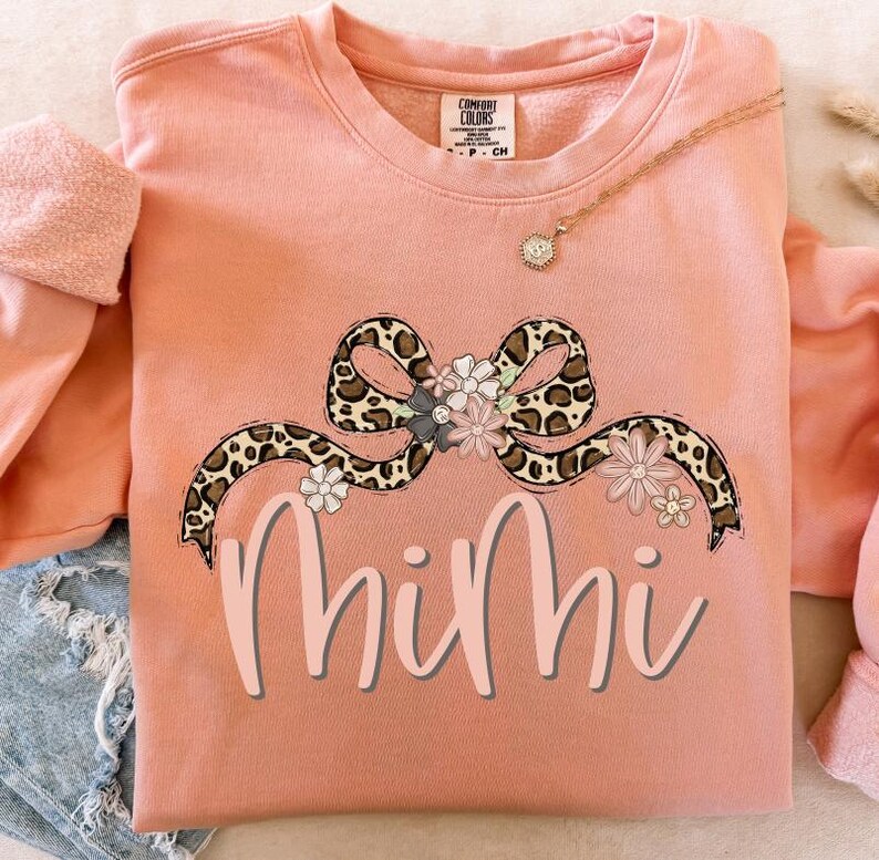 May include: A peach-colored sweatshirt with a leopard print bow and floral accents. The word "Mimi" is written in pink below the bow.