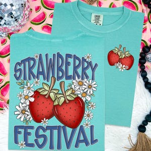 May include: Two turquoise t-shirts with strawberry graphics. One shirt features the text "STRAWBERRY FESTIVAL" with two strawberries and daisies. The other shirt has a smaller strawberry and daisy design. Both shirts are Comfort Colors brand.