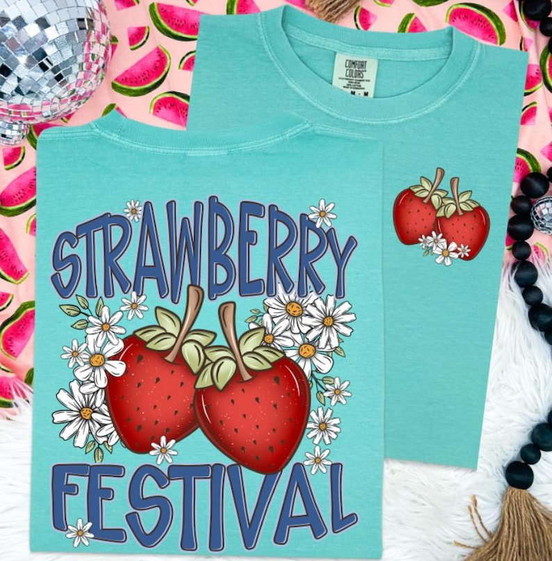 May include: Two turquoise t-shirts with strawberry graphics. One shirt features the text "STRAWBERRY FESTIVAL" with two strawberries and daisies. The other shirt has a smaller strawberry and daisy design. Both shirts are Comfort Colors brand.
