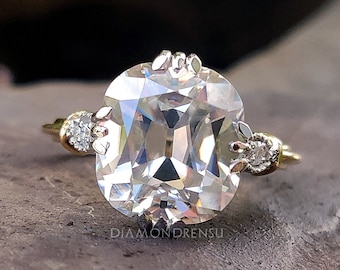 May include: A gold engagement ring with a large, clear, cushion-cut gemstone surrounded by smaller diamonds. The ring is set on a gray, textured surface.