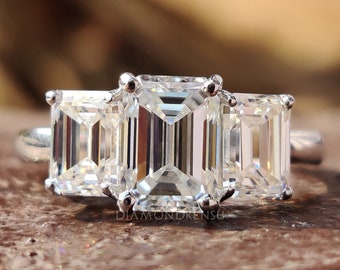 May include: A three-stone emerald cut engagement ring with a silver band. The center stone is larger than the two side stones.