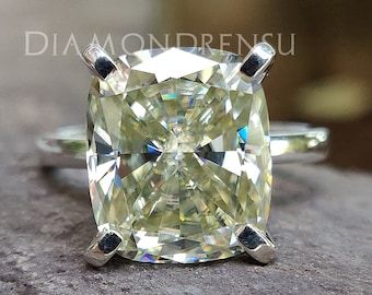May include: A cushion-cut engagement ring with a large, pale yellow diamond set in a silver band. The ring is on a textured, brown surface.