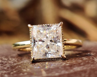 May include: A radiant-cut diamond ring with a gold band. The large, rectangular diamond is set in a gold bezel with a delicate, beaded edge. The ring is displayed on a brown surface, showcasing its brilliance.