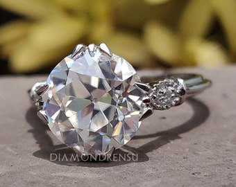 May include: A silver engagement ring with a large, round, clear diamond center stone and two smaller diamonds on the band. The ring is set on a gray, textured surface.
