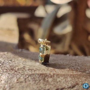 May include: A gold pendant with a blue gemstone set in a square shape. The pendant is on a wooden surface.