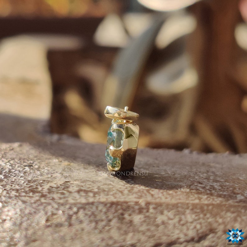 May include: A gold pendant with a blue gemstone set in a square shape. The pendant is on a wooden surface.