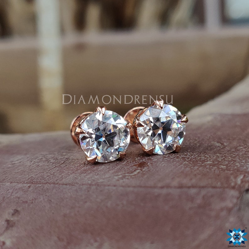 Pode incluir: Um par de brincos redondos de diamantes transparentes engastados em ouro rosa. Os brincos est&atilde;o sobre uma superf&iacute;cie marrom.