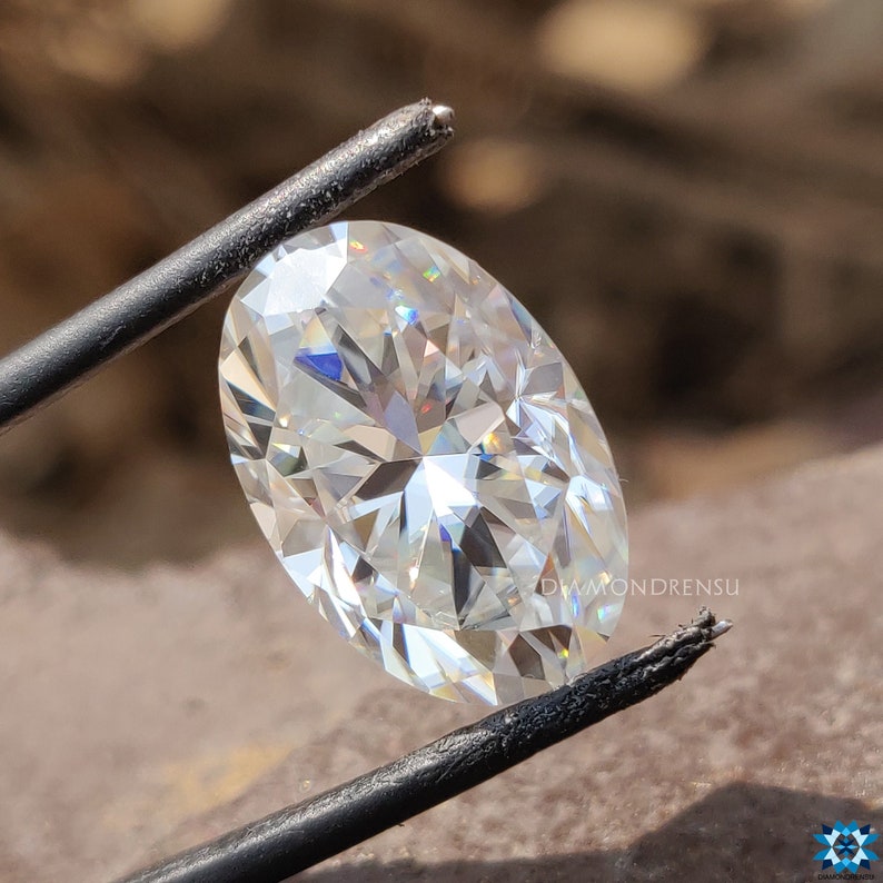 May include: A clear oval-shaped diamond gemstone with facets that reflect light. The diamond is held by a pair of tweezers.