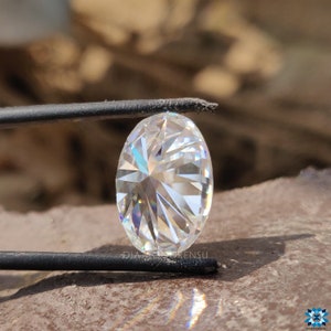 May include: A clear oval-shaped diamond with a brilliant cut, held by tweezers against a brown background.
