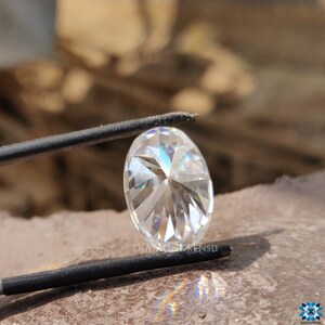 May include: A clear oval-shaped diamond with a starburst pattern in the center. The diamond is held in place by a pair of tweezers.