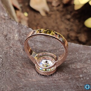 Op de afbeelding: Een verlovingsring van ros&eacute;goud met een grote, heldere, ronde diamant in een bezel-setting. De ring heeft een dunne band en een halo van kleinere diamanten rond de middelste steen.
