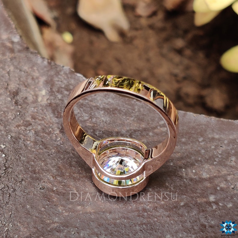 Op de afbeelding: Een verlovingsring van ros&eacute;goud met een grote, heldere, ronde diamant in een bezel-setting. De ring heeft een dunne band en een halo van kleinere diamanten rond de middelste steen.