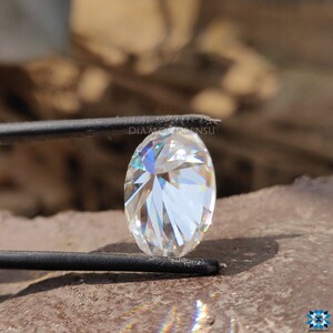 May include: A clear oval-shaped diamond with many facets, held by tweezers. The diamond is reflecting light, creating a rainbow effect.