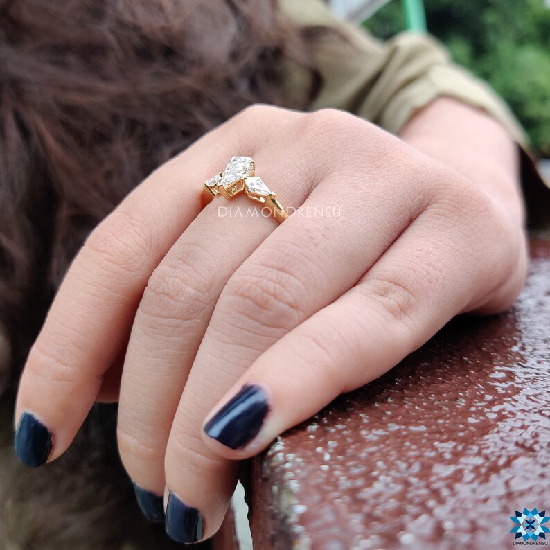 May include: A gold ring with three pear-shaped diamonds set in a prong setting. The ring is on a person's finger.