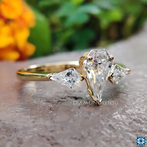 May include: A gold engagement ring with a large pear-shaped diamond and two smaller triangular diamonds on either side. The ring is set on a textured surface.