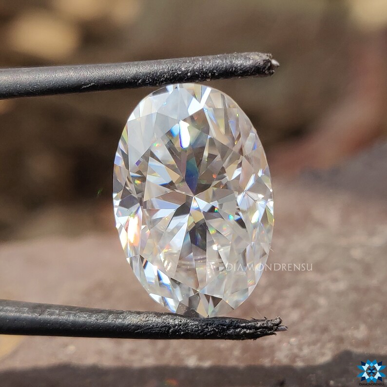 May include: A clear oval-shaped diamond gemstone with a brilliant cut, held by a pair of tweezers. The diamond has a high level of brilliance and sparkle.