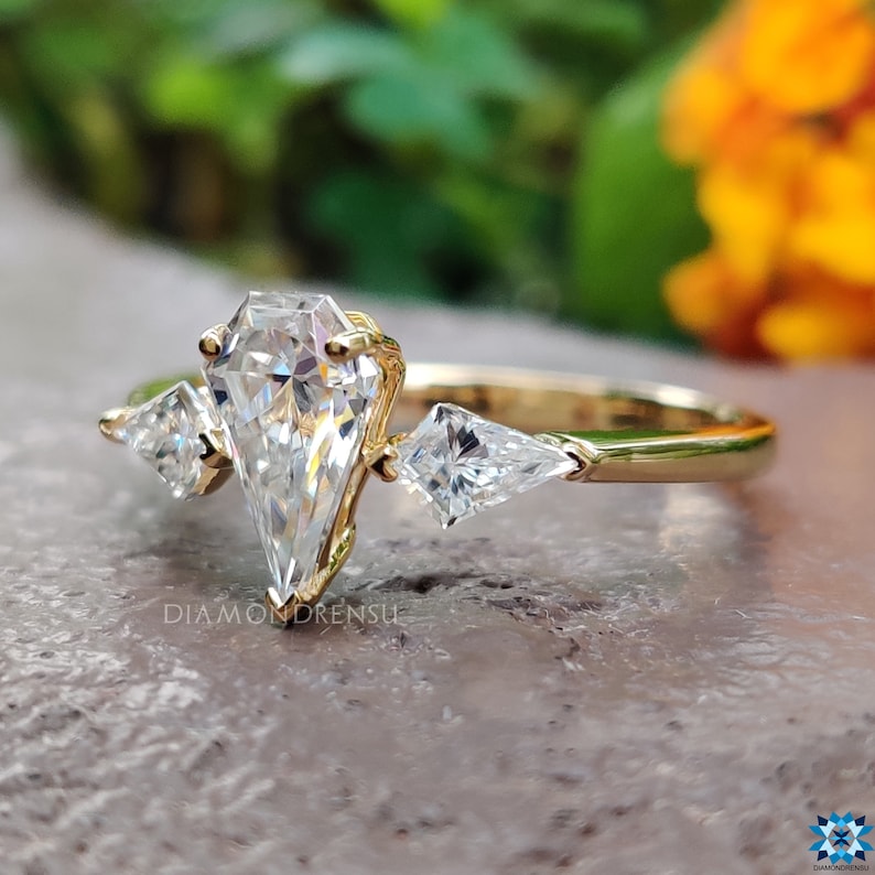 May include: A gold engagement ring with a pear-shaped center stone and two triangular side stones. The ring is set on a thin gold band.