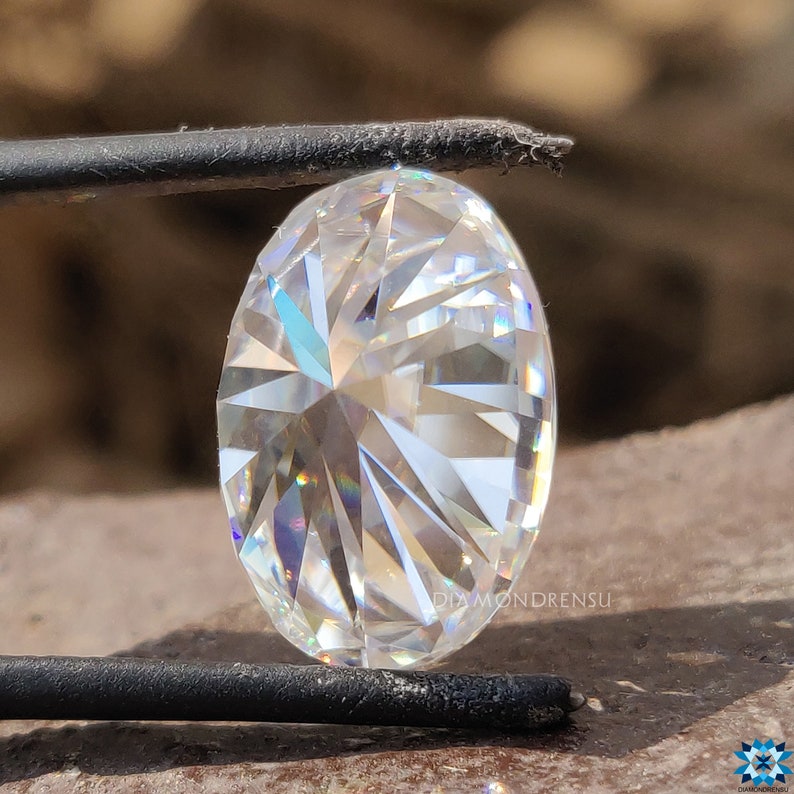 May include: A clear oval-shaped diamond with a brilliant cut, showcasing a starburst pattern of light reflection. The diamond is held by a pair of tweezers.