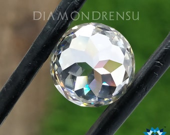 Puede incluir: Una gema redonda, transparente y facetada, con una iridiscencia arcoíris. La gema está sujeta en su lugar por dos barras de metal negro. El texto "DIAMONDRENSU" es visible sobre la gema.