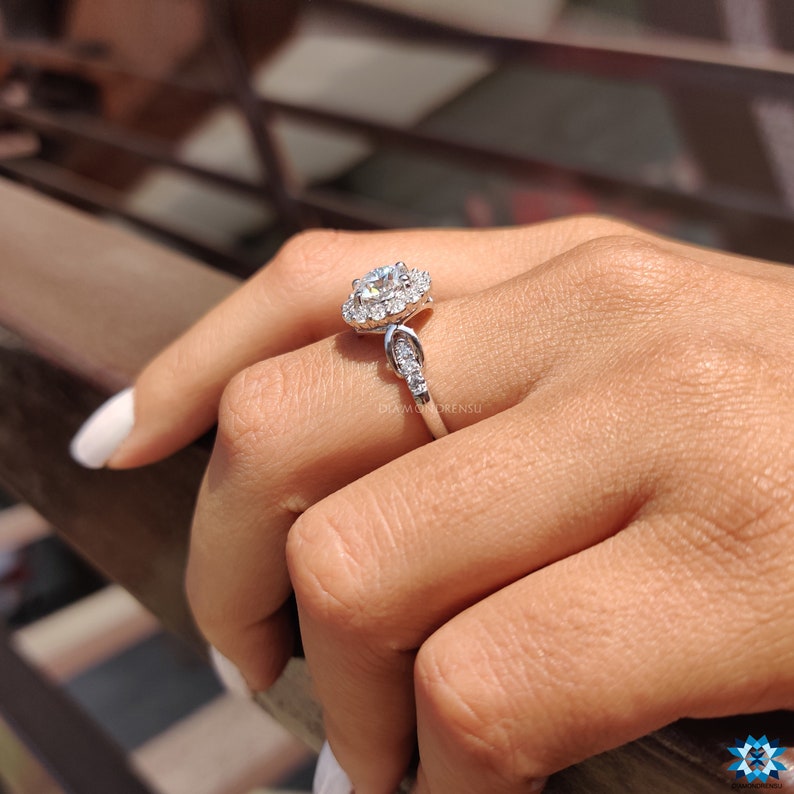 May include: A silver engagement ring featuring a large round centre stone encircled by a halo of smaller stones. The band is embellished with additional smaller stones.