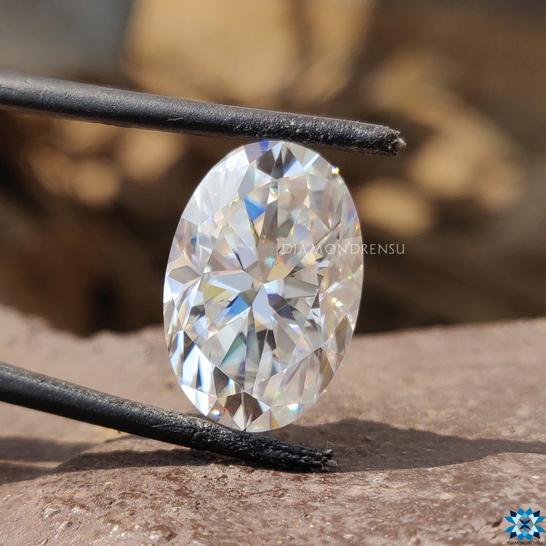 May include: A clear oval-shaped diamond with a brilliant cut, held by tweezers. The diamond is reflecting light, showing its facets.