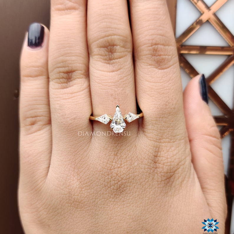 May include: A gold ring with three pear-shaped diamonds. The center diamond is larger than the two side diamonds. The ring is on a person's finger.