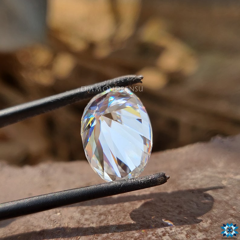 May include: A clear oval-shaped diamond held by tweezers. The diamond has a brilliant cut and reflects light.