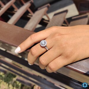 May include: A silver ring featuring a large round diamond encircled by smaller diamonds. The ring is worn on a person's finger.