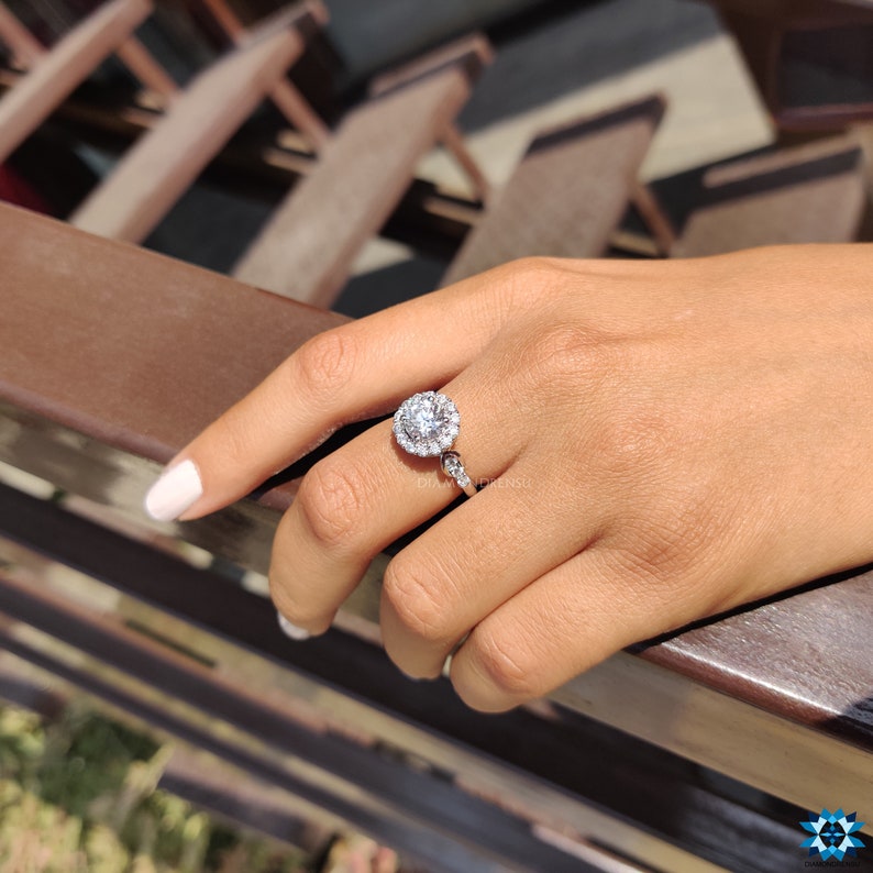 May include: A silver ring featuring a large round diamond encircled by smaller diamonds. The ring is worn on a person's finger.