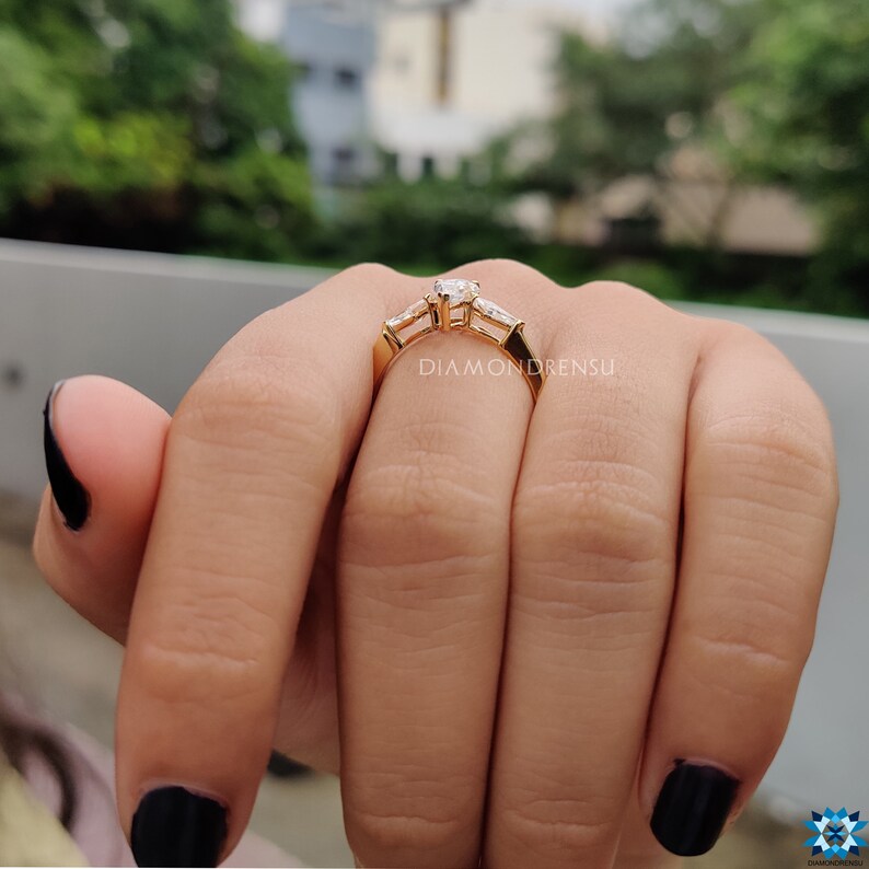 May include: A gold engagement ring with a large round center diamond and two smaller baguette diamonds on either side. The ring is on a person's finger.