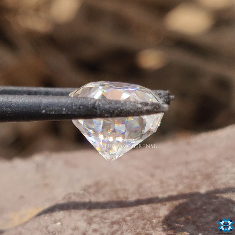 May include: A clear, round, faceted diamond held in tweezers. The diamond has a rainbow-like effect when light shines through it.