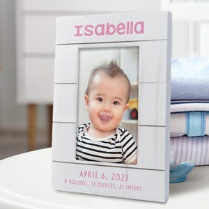 May include: White wooden picture frame with a photo of a baby in a striped shirt. The frame has the name "Isabella" and the date "April 6, 2023" with the baby's birth weight and length.