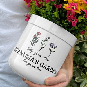 May include: White ceramic flower pot with the text "Grandma's Garden love grows here" and the names "Cathy, James, Elise" printed on it. The pot is filled with pink and yellow flowers.