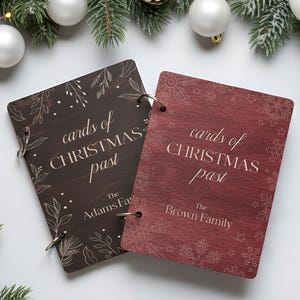 May include: Two wooden Christmas card holders with the text "cards of CHRISTMAS past". One is dark brown with leaf designs, the other is red with snowflake designs. Both have metal ring bindings. The background includes Christmas decorations.