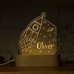May include: A glowing night light featuring a rocket ship design, stars, and the name "Oliver". The light is set on a wooden base and emits a warm, inviting glow, perfect for a child's room. The design includes a planet and stars.