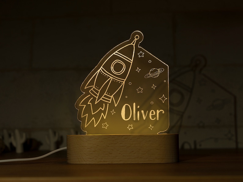 May include: A glowing night light featuring a rocket ship design, stars, and the name "Oliver". The light is set on a wooden base and emits a warm, inviting glow, perfect for a child's room. The design includes a planet and stars.