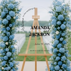 May include: A clear acrylic wedding sign on a wooden easel, with the names "AMANDA and JACKSON" and the date "June 19, 2025". The sign is surrounded by blue floral arrangements. Text reads "Welcome to the wedding of" and "WE ARE SO GLAD YOU ARE HERE".